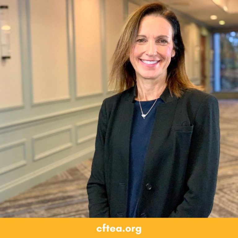 A smiling woman stands in a softly lit hallway with paneled walls and carpeted floors. She has shoulder length brown hair and is wearing a black blazer over a dark blue top with a small necklace. The background is slightly blurred, and a gold bar along the bottom displays cftea.org.