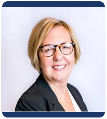 A professional headshot of a woman with short blonde hair and black round glasses, smiling slightly. She is wearing a black blazer over a light top, set against a clean light background. The image is framed with a rounded navy border.