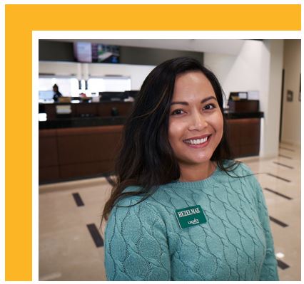 A smiling woman with long dark hair stands inside a bank lobby. She is wearing a teal sweater and a name badge, with teller counters and staff visible in the background. The image is framed by a gold border.