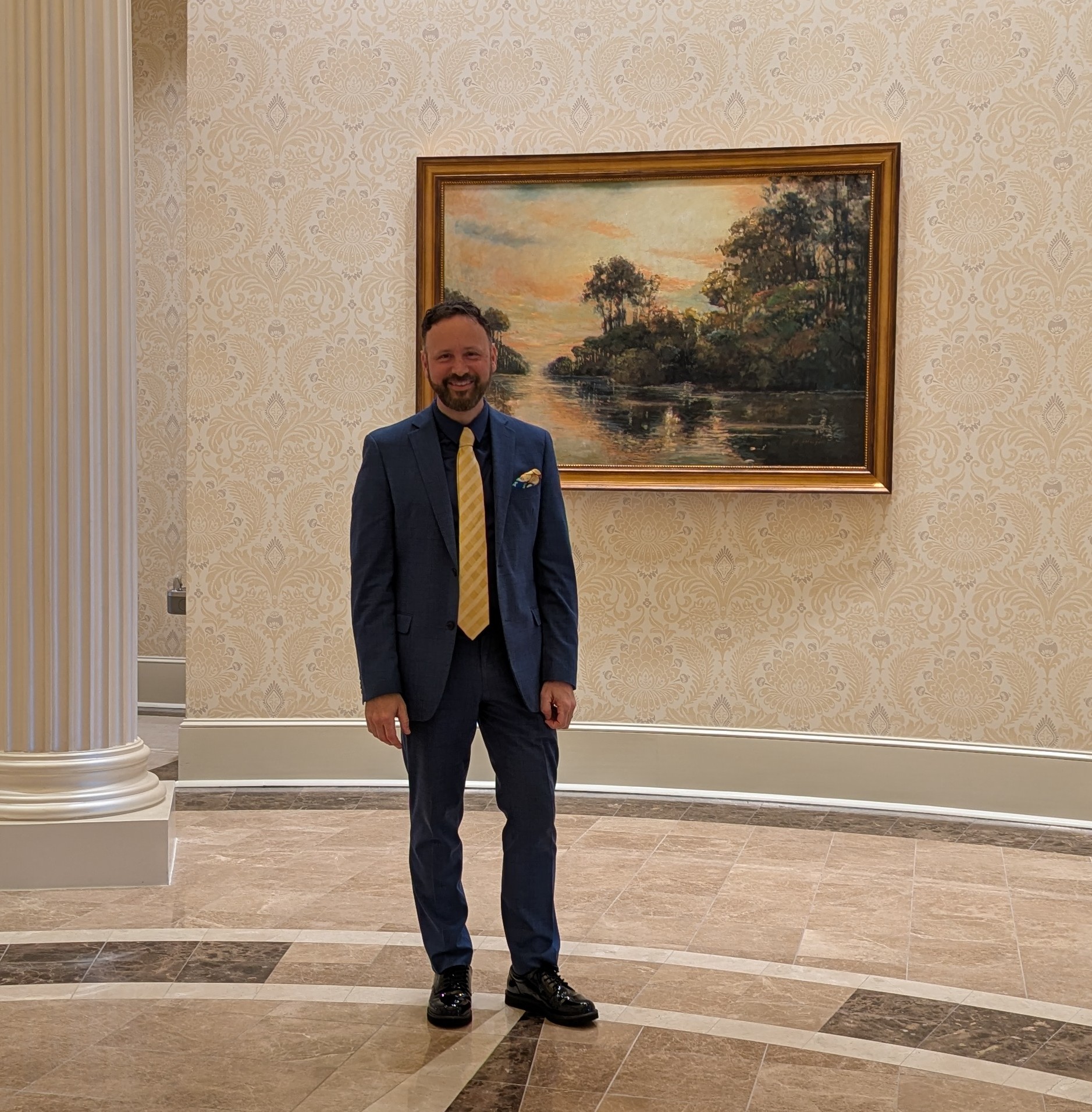 CFTEA Executive Director Andrew Lederer standing in a university building at Towne University, wearing a blue suit and yellow tie, smiling in front of a framed landscape painting at TowneBank.
