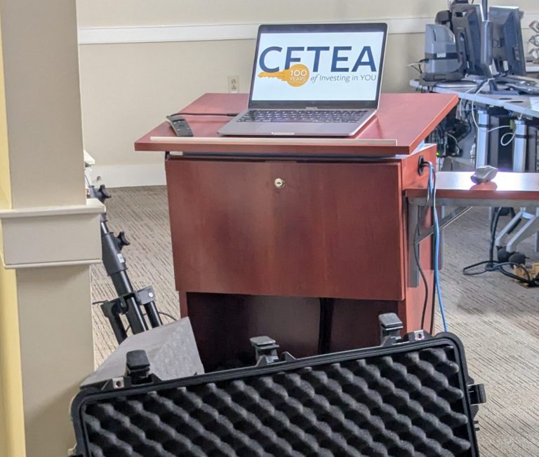 Laptop displaying CFTEA “100 Years of Investing in You” on a podium, with an open equipment case and presentation gear set up in a training room.