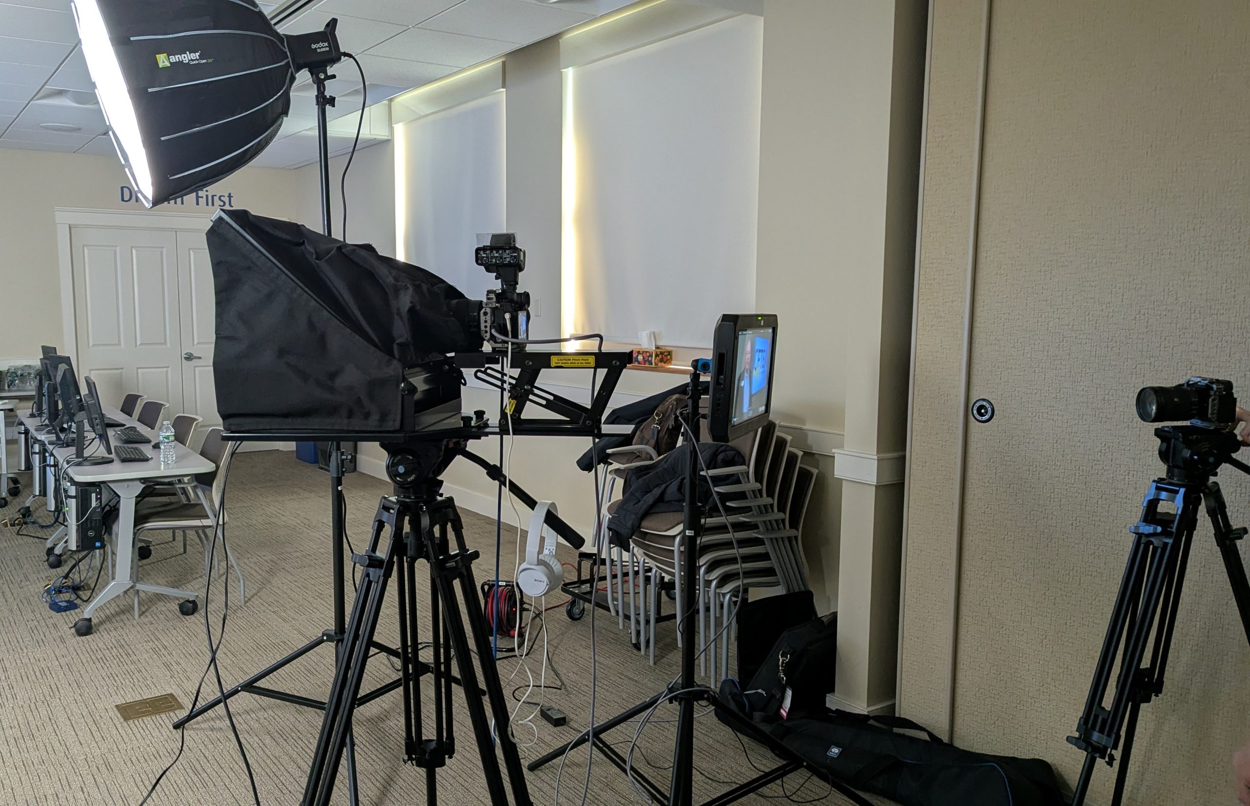 Professional video recording setup in a training room with cameras on tripods, lighting equipment, teleprompter, and computer stations prepared for filming or virtual instruction.