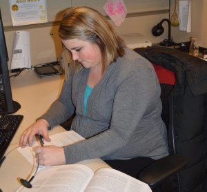 A professional sits at a desk in a modern office workspace, focused on completing coursework while referencing an open textbook. A computer monitor and keyboard sit nearby, highlighting a blend of workplace responsibilities and continuing education. The clean, uncluttered office setting emphasizes concentration, learning, and professional development.